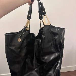 Svart axelväska med gulddetaljer från Dknk - Snygg svart axelväska i syntetmaterial med eleganta gulddetaljer. Väskan har en praktisk dragkedja och en dekorativ medaljong. Perfekt för att bära med sig det nödvändigaste med stil.