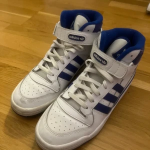 Vita och blå sneakers från Adidas - Snygga vita och blå sneakers från Adidas med klassiska ränder och snörning. Skorna har en hög modell med kardborreband för extra stöd. Perfekta för en sportig look. Köpta 2022 och använda kanske 3ggr max, men gör rent dem vid köp. Storlek 41 1/2, nypris 1450kr