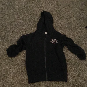 Mörkblå hoodie från Tommy Hilfiger - Snygg svart hoodie från Tommy Hilfiger med dragkedja och logga på bröstet. Perfekt för en casual look med sin klassiska design och bekväma passform.
