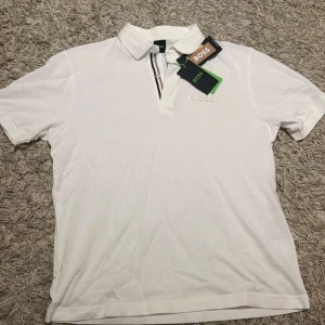 Vit kortärmad pikétröja från Boss - Snygg vit pikétröja från Boss med korta ärmar och klassisk krage. Tröjan har en diskret logga på bröstet och en stilren randdetalj på baksidan. Perfekt för en stilren och avslappnad look.