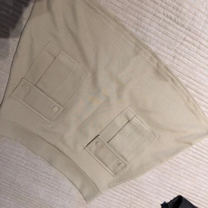 Beige kjol med fickdetaljer - Snygg beige kjol med två stora fickor framtill och en diskret dragkedja i sidan. Perfekt för en stilren look. Köpt i somras använd 1 gång.