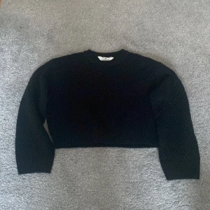 Svart stickad tröja från H&M - Säljer en svart stickad tröja från H&M med ett hålmönster. Tröjan har långa ärmar och en croppad passform, perfekt för lager på lager-stil. Den är både stilren och bekväm, idealisk för kyligare dagar. Man kan ha en svart långärmad tröja under.