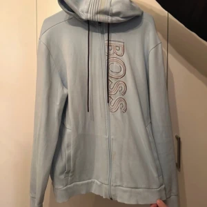 BOSS hoodie - Säljer en stilren ljusblå hoodie från BOSS med dragkedja och justerbar huva. Tröjan har en broderad logotyp på bröstet och ribbade muddar vid ärmslut och nederkant. Perfekt för en avslappnad stil.