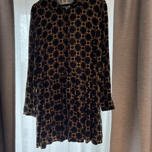 Svart mönstrad klänning från Amisu/New Yorker - Snygg svart klänning från Amisu med ett kedjemönster i guld. Klänningen har långa ärmar och en klassisk krage. Perfekt för en stilren look.