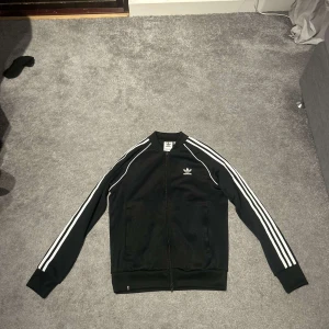 Adlads zip  - Säljer en klassisk svart träningsjacka från Adidas med vita ränder längs ärmarna. Jackan har dragkedja framtill och är långärmad med ribbade muddar. Perfekt för en sportig look.