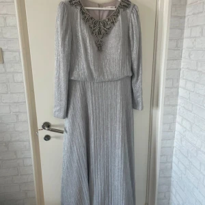 Silverfärgad långklänning med broderad detalj - Elegant silverfärgad långklänning med vackra broderade detaljer vid halsringningen. Klänningen har långa ärmar och ett subtilt glittrande material som ger en lyxig känsla. Den har dessutom en built- in bh. Perfekt för speciella tillfällen. Strl 44 MEN passar strl 40 och 42. Använt en gång så den är fortfarande i väldigt bra skick.