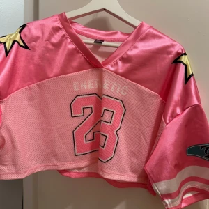 Rosa croppad tröja från Shein  - Snygg rosa croppad tröja från Shein med nummer 23 tryckt på framsidan. Tröjan har en v-ringad hals och stjärndetaljer på axlarna. Perfekt för en sportig look! MESH. 