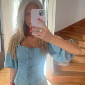 Ljusblå jeanskorsett - Trendig ljusblå korsett i jeanstyg med puffiga kortärmar och knappar framtill. Perfekt för en stilren look.