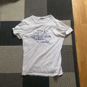 Vit t-shirt från Calvin Klein Jeans - Säljer en klassisk vit t-shirt från Calvin Klein Jeans med det ikoniska CK-logotypen tryckt i blått på framsidan. T-shirten är kortärmad och gjord i mjukt bomullsmaterial. 