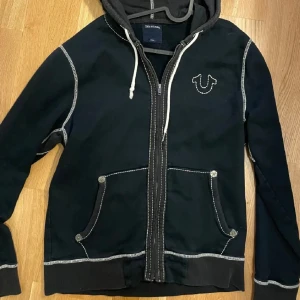 Svart hoodie med dragkedja från True Religion - Snygg svart hoodie från True Religion med dragkedja och broderad logga på bröstet. Den har kontrasterande vita sömmar och en praktisk huva med snörning. Perfekt för en avslappnad stil. Står L men passar S-M. Har tappat pyttelite färg i tvätten.