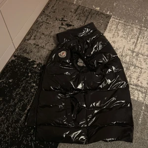 Svart pufferjacka från Moncler - Snygg svart pufferjacka från Moncler med glansig finish. Jackan har en klassisk design med Moncler-logga på bröstet. Perfekt för kyligare dagar med sin isolerande stil.