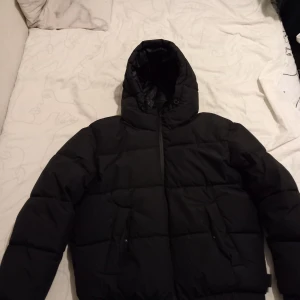 Svart pufferjacka från Bershka - Säljer en svart pufferjacka från Bershka med huva och dragkedja. Jackan är perfekt för kalla dagar och har en stilren design. Inget fel med den