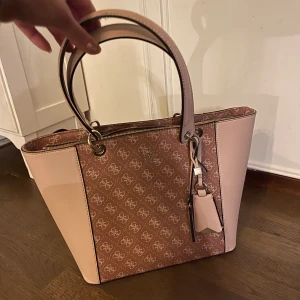 Rosa och beige axelväska från Guess - Snygg axelväska från Guess i rosa och beige med monogrammönster. Väskan har dubbla handtag och en praktisk dragkedja upptill. Perfekt för att bära dina dagliga nödvändigheter med stil. Förutom slitage på handtag (3 skrapmärken) är väskan i skick som ny. 
