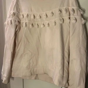 Beige tröja med tofsdetaljer - Säljer en beige tröja med unika tofsdetaljer längs ärmarna och bröstet. Tröjan har en avslappnad passform och långa ärmar, perfekt för en mysig dag.