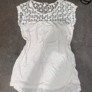 Vit blus med spetsdetaljer - Säljer en söt vit blus med vackra spetsdetaljer upptill. Blusen är ärmlös och har en rund halsringning. Perfekt för en somrig look!