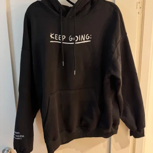 Svart hoodie med motiverande text - Säljer en svart hoodie med texten 'KEEP GOING:' på framsidan och '100 REASONS TO STAY ALIVE:' på baksidan. Den har en känguruficka och justerbar huva med snören. Perfekt för att hålla sig varm och motiverad under kyliga dagar. Passar både för vardag och mysiga hemmakvällar.