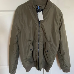 Olivgrön bomberjacka från H&M - Säljer en snygg olivgrön bomberjacka från H&M i mycket bra skick. Jackan har en dragkedja framtill och två fickor med knappar. Perfekt för höst och vår, med ribbade muddar vid ärmslut och krage. Passar både till vardags och fest!