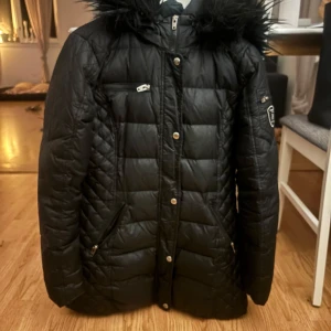 Svart dunjacka med pälshuva - Säljer en snygg svart dunjacka med pälshuva. Jackan har dragkedja och knappar framtill, samt praktiska fickor med dragkedja. Perfekt för kalla vinterdagar! 🖤