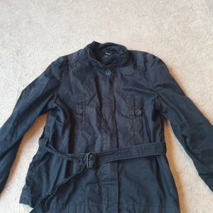 Svart jacka från Filippa K - Säljer en stilren svart jacka från Filippa K i bomull. Den har långa ärmar och ett bälte i midjan för en snygg passform. Perfekt för höst och vår! Jackan har knappar framtill och en hög krage. Fint skick, inga hål eller dylikt men något urtvättad, samt fläck på ena ärmen, se bild 🌸