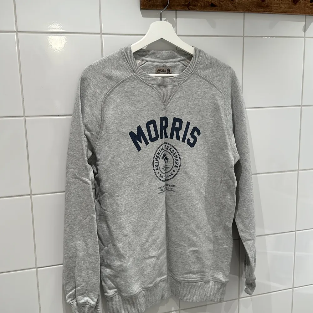 Säljer en snygg grå sweatshirt från Morris i storlek M. Tröjan har ett tryck med Morris-loggan i marinblått på framsidan. Den är i bra skick och perfekt för både höst och vår. Mjuk och bekväm med långa ärmar och ribbade muddar.. Tröjor & Koftor.