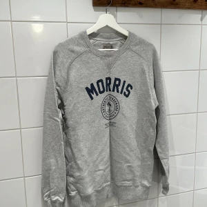 Grå sweatshirt från Morris - Säljer en snygg grå sweatshirt från Morris i storlek M. Tröjan har ett tryck med Morris-loggan i marinblått på framsidan. Den är i bra skick och perfekt för både höst och vår. Mjuk och bekväm med långa ärmar och ribbade muddar.