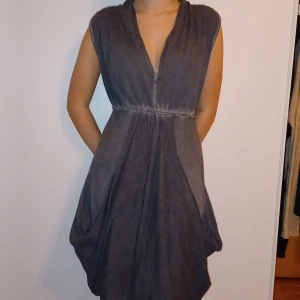 Grå ärmlös klänning - Säljer en stilren grå klänning i ett mjukt material. Den är ärmlös med en v-ringad hals och har en knytning i ryggen för en justerbar passform. Perfekt för både vardag och festliga tillfällen!