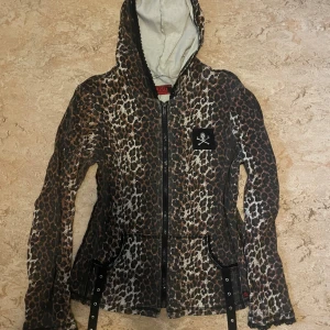 Leopardmönstrad jacka med huva från Tripp - Säljer en cool leopardmönstrad jacka från Tripp med huva och dragkedja. Den har ett dödskallemärke på bröstet och snygga detaljer som svarta remmar med nitar. Perfekt för höst och vår när du vill sticka ut lite extra!