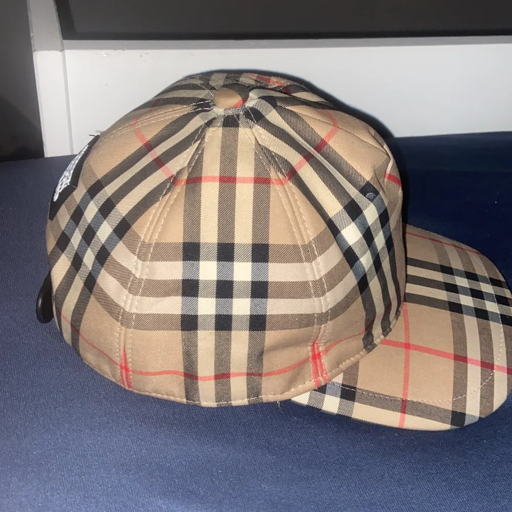 Snygg rutig keps från Burberry i klassiskt beige, svart och rött mönster. Perfekt för att ge din outfit en stilren touch. Justerbar passform baktill med en svart rem. Passar både till vardags och festliga tillfällen.. Accessoarer.