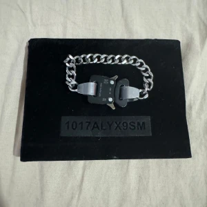 Silverfärgat armband från 1017 ALYX 9SM - Säljer ett stilrent och modernt armband från 1017 ALYX 9SM. Armbandet är silverfärgat med en robust kedja och en unik svart spänndetalj. Perfekt för att ge din outfit en edgy touch. Kommer med originalförpackning.