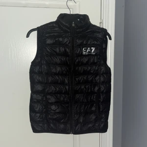 Svart dunväst från EA7 - Säljer en svart dunväst från EA7 i bra skick. Västen har en dragkedja framtill och är quiltad för extra värme. Perfekt för höst och vår. Det finns en liten skada på framsidan, men den påverkar inte funktionen. Passar bra till både vardag och fest. OBS enda deffekten är ett litet streck hål i den men syns inte om man inte kollar noga 