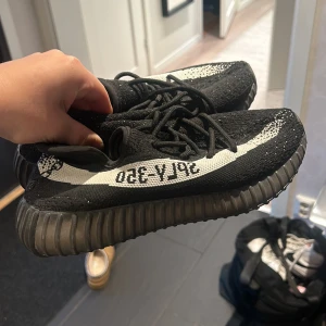 Svarta Adidas Yeezy Boost 350 V2 - Säljer ett par svarta Adidas Yeezy Boost 350 V2 i mycket bra skick. De har en snygg svart och vit design med den ikoniska 'SPLY-350' texten på sidan. Skorna har en bekväm passform och är perfekta för både vardag och fest. De är i storlek herr 42 och har en robust sula för extra komfort.