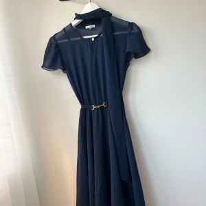 Mörkblå klänning från Ida Sjöstedt - Elegant mörkblå klänning från Ida Sjöstedt i storlek 34. Klänningen har korta ärmar och ett stilrent bälte i midjan som ger en fin siluett. Perfekt för både fest och formella tillfällen. Materialet känns lätt och luftigt, vilket gör den bekväm att bära hela dagen.