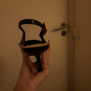 Svarta klackskor med spetsig tå - Säljer ett par eleganta svarta klackskor med spetsig tå och en stilren design. Skorna har en smal rem med gulddetalj och är perfekta för festliga tillfällen. De är i mycket bra skick och redo att användas för att lyfta din outfit.