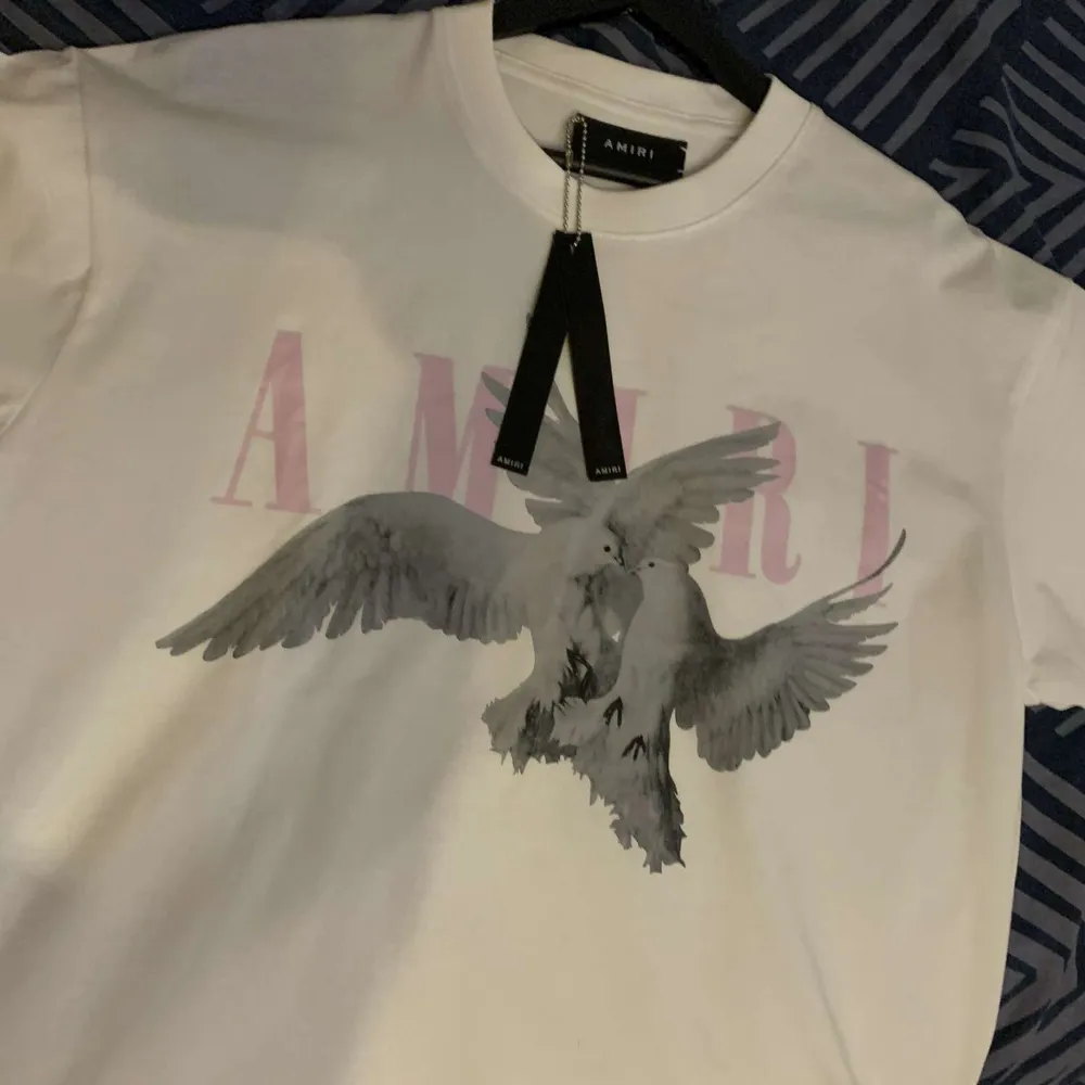 Säljer en snygg vit t-shirt från Amiri med ett coolt duvmotiv och rosa text. Perfekt för en avslappnad stil. T-shirten är i nytt skick och passar perfekt till både jeans och shorts. Skön och stilren design!. T-shirts.