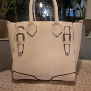 Vit handväska med spänndetaljer - Säljer en stilren vit handväska med snygga spänndetaljer i silver. Väskan har två handtag och en modern design som passar perfekt för både vardag och fest. Den är rymlig och har en elegant look. Perfekt för att lyfta vilken outfit som helst!