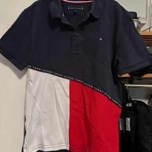 Flerfärgad pikétröja från Tommy Hilfiger - Säljer en snygg pikétröja från Tommy Hilfiger i blått, vitt och rött. Tröjan har korta ärmar och en klassisk krage med knappar. Den är gjord av 100% bomull och har en broderad logga på bröstet. Perfekt för en avslappnad men stilren look!