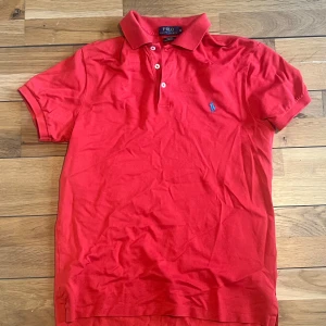 Röd pikétröja från Polo Ralph Lauren - Säljer en klassisk röd pikétröja från Polo Ralph Lauren. Den har en broderad logga på bröstet och knappar vid kragen. Perfekt för en avslappnad men stilren look. Passar både till vardags och fest!