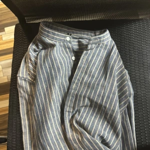 Randig skjorta i blått och vitt - Säljer en snygg randig skjorta i blått och vitt. Skjortan har en klassisk design med knappar framtill och är perfekt för både vardag och fest. Materialet känns bekvämt och luftigt, vilket gör den idealisk för vår och sommar. Passar bra till jeans eller chinos för en avslappnad look.