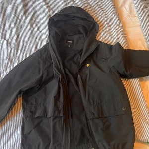 Svart jacka från Lyle & Scott Junior - Säljer en svart jacka från Lyle & Scott Junior i mycket bra skick. Jackan har en stilren design med dragkedja och huva, perfekt för höst och vår. Den är tillverkad i ett syntetmaterial och har ett litet broderat emblem på bröstet. Passar perfekt för både skola och fritid!