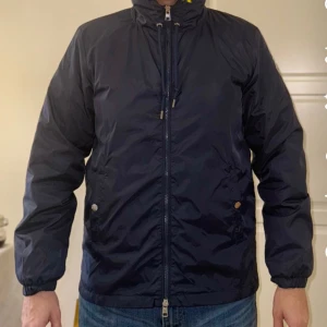 Svart vindjacka från Moncler - Säljer en snygg svart vindjacka från Moncler i mycket bra skick. Jackan har en dragkedja framtill och justerbar huva med röd och vit kant. Perfekt för höst och vår! Den har långa ärmar och två fickor med knappar. Moncler-loggan är broderad på bröstet.