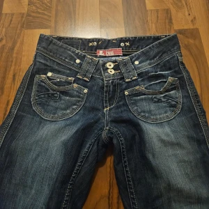 Mörkblå bootcut jeans med unika fickdetaljer - Säljer ett par mörkblå bootcut low rise jeans från vinted som tyvärr var för små för mig. Bra skick, coola fickdetaljer och dubbla knappar i midjan. Skedjan är lite sönder men fungerar fortfarande och går att fixa.                       Måtten: Midjemått tvärs över: 38cm Lårvidd: 47 cm Innrebensmått: 80 cm Yttrebensmått: 98 cm 