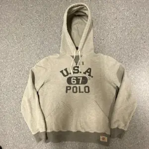 Säljer en snygg grå hoodie från Polo Ralph Lauren med trycket 'U.S.A. 67 POLO' på framsidan. Den har en klassisk känguruficka och justerbar huva med snören. Perfekt för en avslappnad stil eller kyliga dagar. 🐴