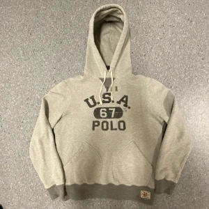 Grå unik  hoodie från Polo Ralph Lauren - Säljer en snygg grå hoodie från Polo Ralph Lauren med trycket 'U.S.A. 67 POLO' på framsidan. Den har en klassisk känguruficka och justerbar huva med snören. Perfekt för en avslappnad stil eller kyliga dagar. 🐴