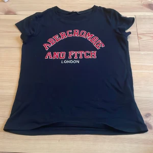 Mörkblå t-shirt från Abercrombie & Fitch - Säljer en mörk blå t-shirt från Abercrombie & Fitch i storlek XS. Den har ett coolt tryck med texten 'Abercrombie and Fitch London' i rött och vitt på framsidan. På ärmen finns en broderad brittisk flagga och A&F-logga. Perfekt för vardagsbruk!