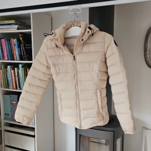Beige dunjacka från Napapijri - Säljer en snygg beige dunjacka från Napapijri. Den har en dragkedja framtill och en mysig huva. Perfekt för kyliga dagar och håller dig varm och stilren. Jackan är i mycket bra skick och passar perfekt för höst och vinter.