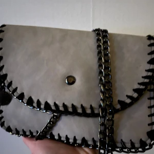 Grå axelväska med kedjedetaljer - Snygg grå axelväska i syntetmaterial med coola kedjedetaljer och svart kant. Väskan har en praktisk storlek och stängs med en knapp. 