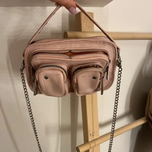 Rosa axelväska från Núnoo - Säljer en superfin rosa axelväska från Núnoo i skinn. Den har flera praktiska fickor med dragkedjor, två axelband ingår och även ett litet hänglås som man kan låsa olika fack med. Perfekt för både vardag och fest! 💖