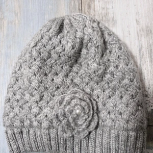 Grå stickad mössa från KappAhl - Superfin grå stickad mössa från KappAhl i one size. Den har ett snyggt flätmönster och en söt blomdetalj med en pärla framtill. Perfekt för kalla höst- och vinterdagar. Materialet är 100% akryl med ett mjukt polyesterfoder för extra värme.