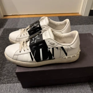 Valentino sneakers - Säljer ett par snygga Valentino sneakers i vitt skinn med svarta detaljer och en stjärna på framsidan. Skorna har en bred rem med VLTN-logga och är i mycket bra skick.