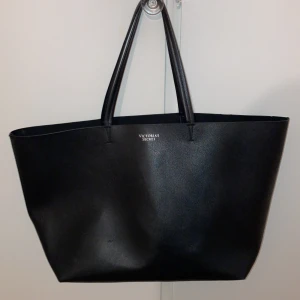 Svart Victoria's Secret totebag - Snygg och rymlig svart totebag från Victoria's Secret. Perfekt för en dag på stan eller som en stilren vardagsväska. Tillverkad i syntetmaterial med två långa handtag för enkel bärning. Diskret logga i silver på framsidan ger en elegant touch. Pris kan diskuteras! Köpt för 500kr men säljer för 150kr